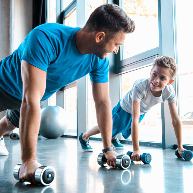 Dad and son at the gym in a plank position smiling at each other while working out together