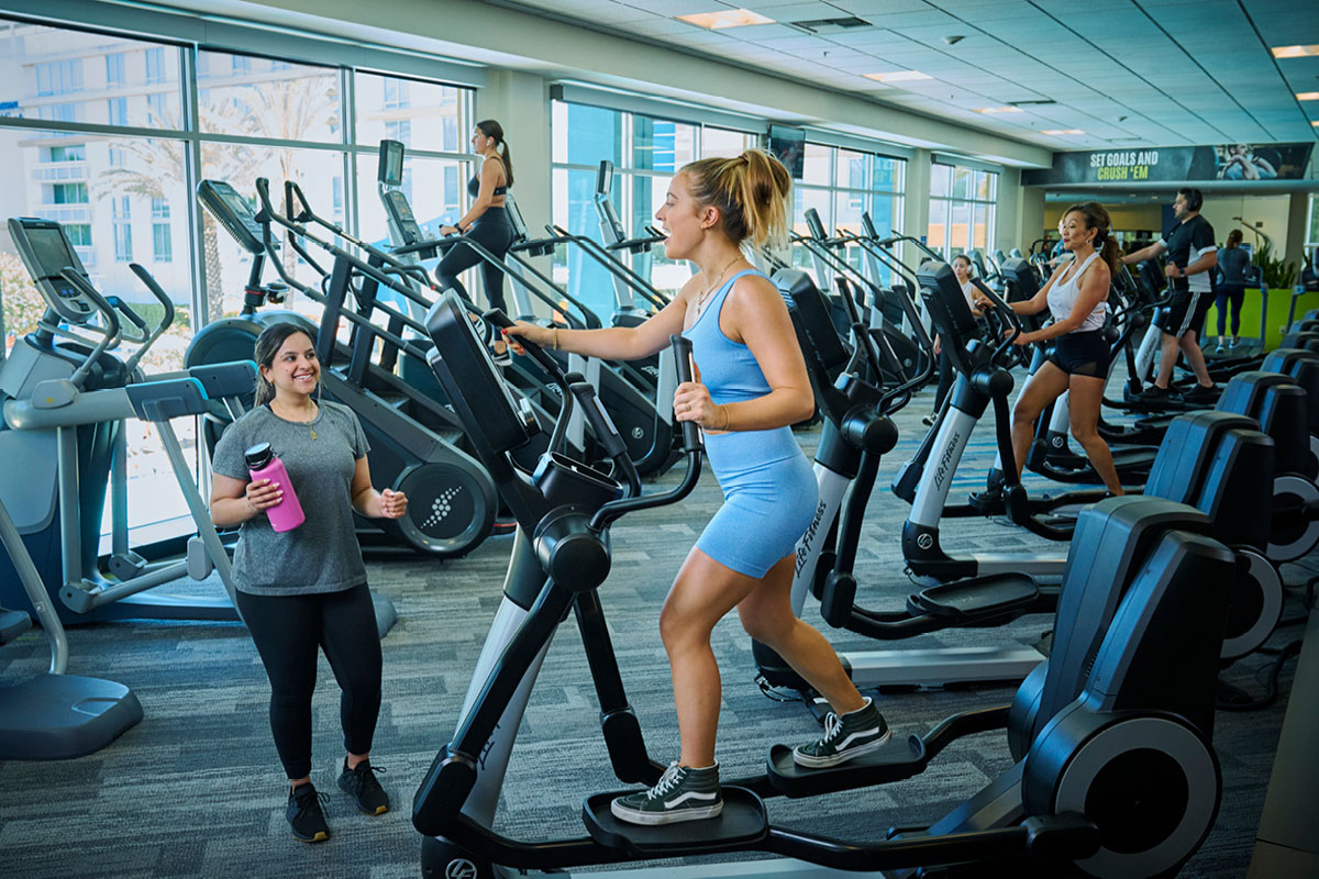 Woman on the fitness floor working out on an elliptical trainer chatting with a friend walking by