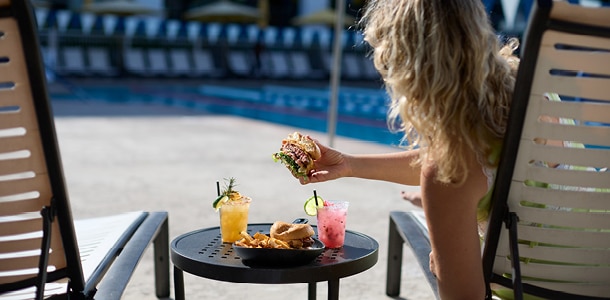 A woman enjoying a burger and cocktails while lounging by the outdoor pool