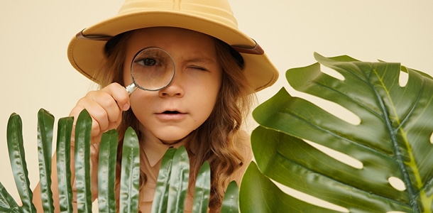 A little girl in safari attire looking through the bushes with a magnifying glass